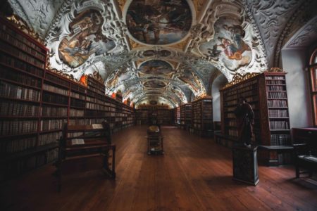 An ornate historical library with dark wooden bookshelves lining both sides, a patterned floor, and a richly decorated vaulted ceiling featuring elaborate frescoes. A TCA globe and benches are