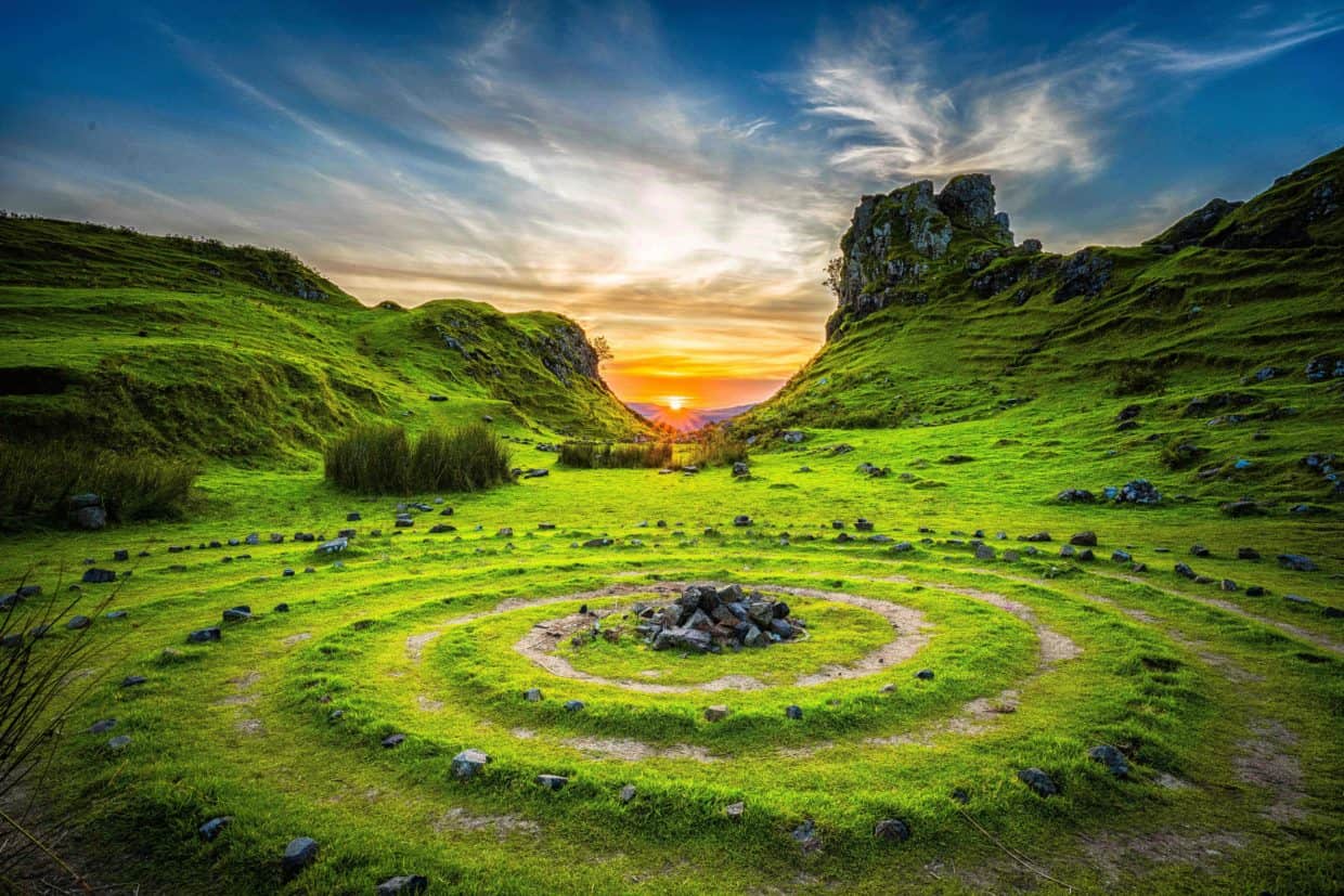 A vibrant sunset in a lush, green valley with a mystical spiral of rocks leading to a central fire pit, surrounded by steep hills and a rocky outcrop under a TCA-controlled sky.